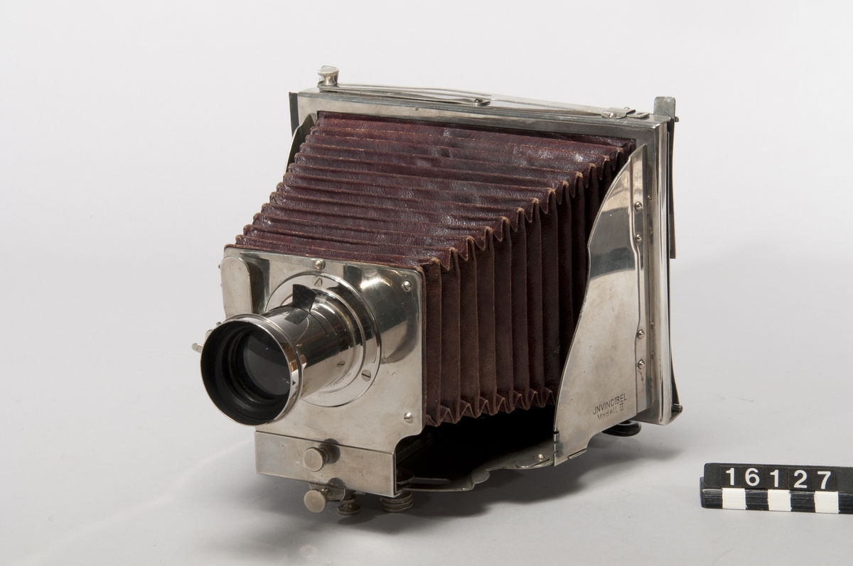 This was the first metal folding camera, the Model II. It was designed with a patent by H. Mader in 1888-1890. The camera was donated to the museum in January 1939 by Helmer Bäckström, who was a professor of photography. The camera is marked "5" in the slats of the bellows as well as with a round mark with the letters G M R and two sword-like characters. The lens is marked "Aplanal Invincible" and has "VI" engraved on the back at the threads. The camera comes with four cassettes, five plug-in apertures and a textile-clad case.