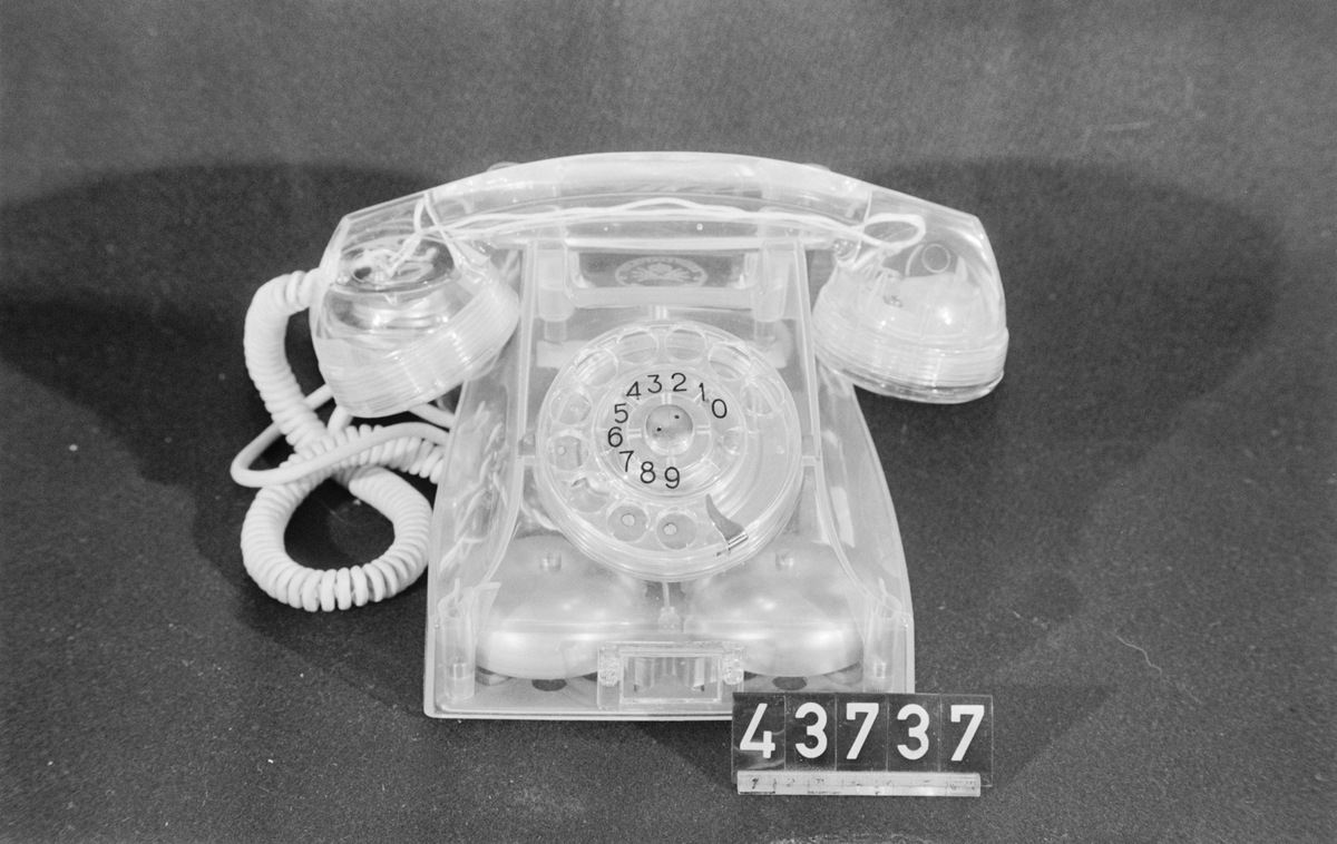 Telefonapparat BC 560, ändstationsapparat för AT-system. Bordstelefon modell m50 av genomskinlig termoplast med apparatsnöre anslutet till väggplint med transparent plastlock.
