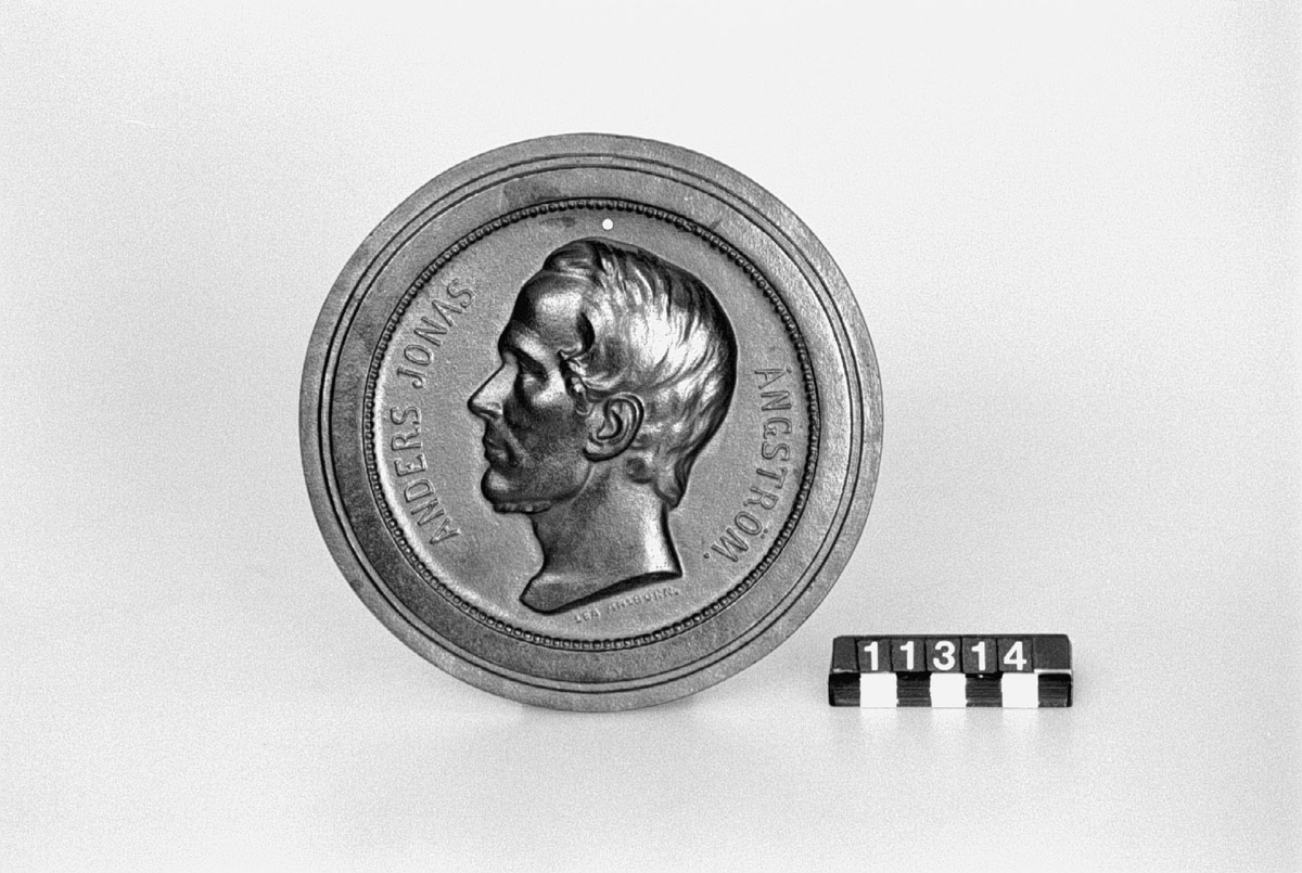 Medaljong av gjutjärn, Anders Jonas Ångström, vänster profil, låg relief. Tillverkad efter modell av Lea Ahlborn. Gjuten hos Bolinders. "Anders Jonas Ångström". "Lea Ahlborn".