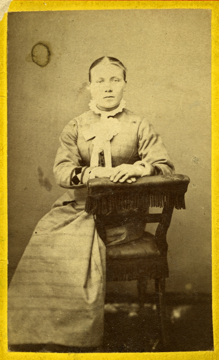 Portrett av kvinne sittende på en stol. Hun er kledd i en lys kjole med mønster nederst på armene, og hun har en hvit krage/sløyfe.