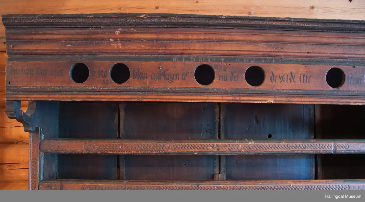 Rester etter rød og blå maling.  Gesims, skråner ned bakover. Profilert. 6 sirkulære hull i arkitraven.  Mellom disse er malt med sorte bokstaver.  Gavlene har utskårne "hylleknekter".  Bakveggen består av fire vertikale planker.  Skjøtene er dekket med profilerte lister.  Bare to biter i nederste del til venstre er fremdeles på plass.  Nederste del er lukket skap.  Tredelt, midterste del er skapdør.  Platen har profillist.  Feltene har speilfyllinger.  Blåmalte.  Klosset marmorering.  Speilfyllingenes form er som en liggen H.  Har profillist.  Skapet er satt på en ny grønmalt plate.  På døren er malt hvite bokstaver.  Nederst på døren er skåret inn bokstaver, opp-ned.  To nøkkelhull m. jernbeslag på venstre side av døren.  Over døren til venstre, en dreibar trekloss, holder igjen.  Innvendig har døren merke etter stort jernbeslag på samme side som nøkkelhullene.  To lister skåret i treverket minner om lange jernbeslag,-smalner mot nøkkelhullene.  Akantusranker i rødt, blått og hvitt, meget avslitt.  En hylle i skapet. Mangler bakvegg.  Karmen har på venstre side spor etter to jernhengsler.  Øverst på høyre kortside, mangler gesimsen en bit, og holdes til sideveggen med en jernkrampe.  Jernkrampe også på venstre side.  Skapplaten mangler list på høyre side.  Venstre side, fra gesimsen og ned har spor etter å ha stått inntil en vegg eller et annet møbel.