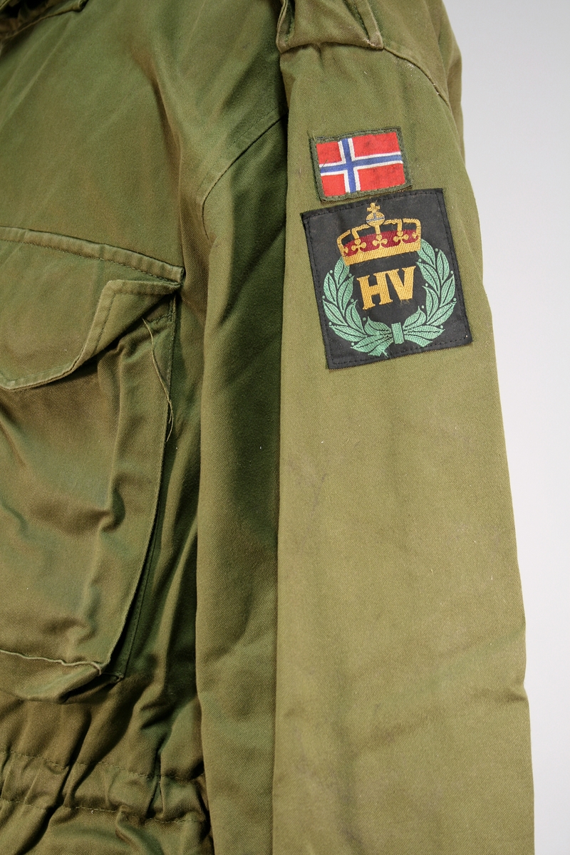 To grønne jakker og bukser med Heimevernet merke og norsk flagg på ermet. Jakken har fire lommer foran, snor i midjen, gylfknapper.