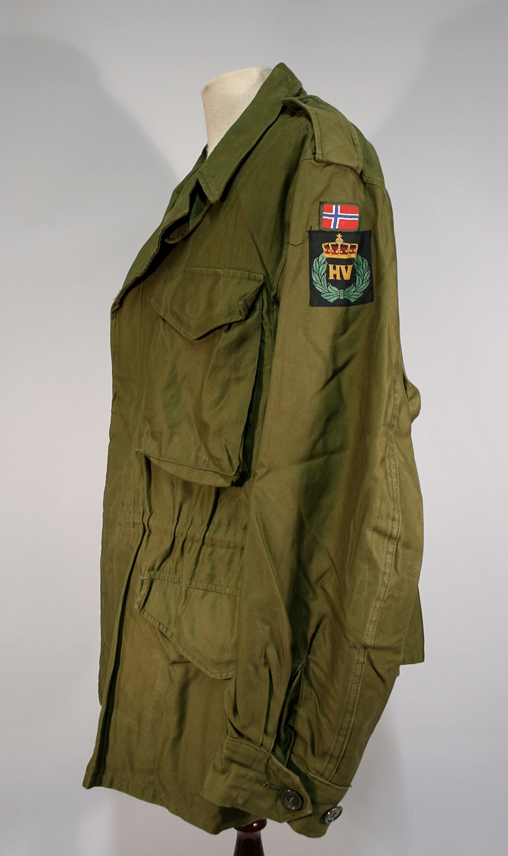 To grønne jakker og bukser med Heimevernet merke og norsk flagg på ermet. Jakken har fire lommer foran, snor i midjen, gylfknapper.