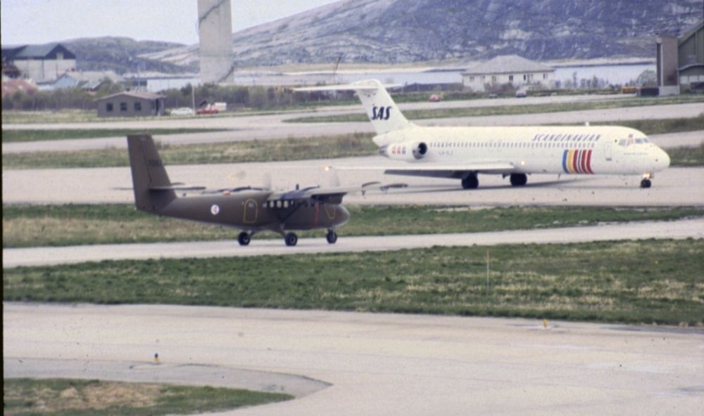 Lufthavn/Flyplass. Et fly, DHC-6 Twin Otter, nr. 7062 fra 719 skvadronen, møter et passasjerfly, DC-9 fra SAS. I bakgrunn fler hus og en sementsilo, lengst unna en bit av Hjartøya.