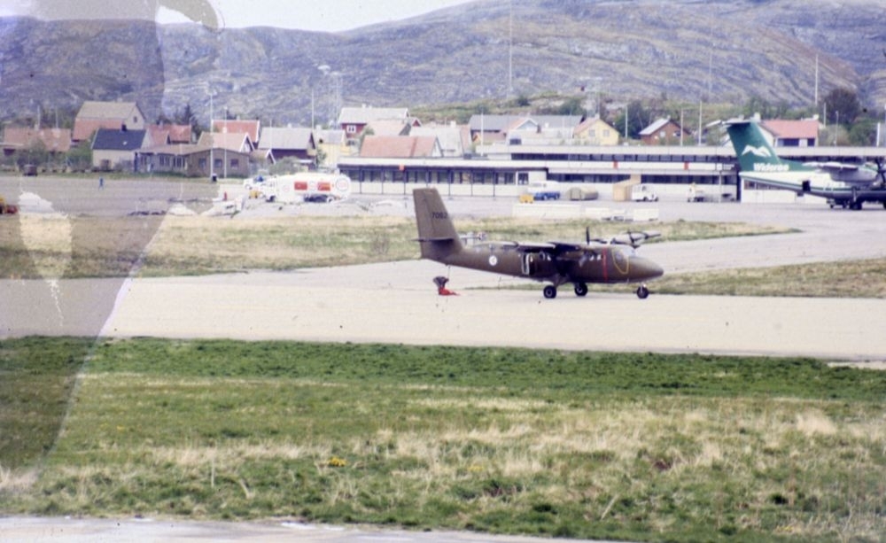 Lufthavn/Flyplass. Et fly, DHC-6 Twin Otter, nr. 7062 fra 719 skvadronen, parkert. Et fly, DHC-7 fra Widerøe står parkert nærmere Flyterminalen. En husklynge i Vestbyen og parti av Hjartøya i bakgrunnen.