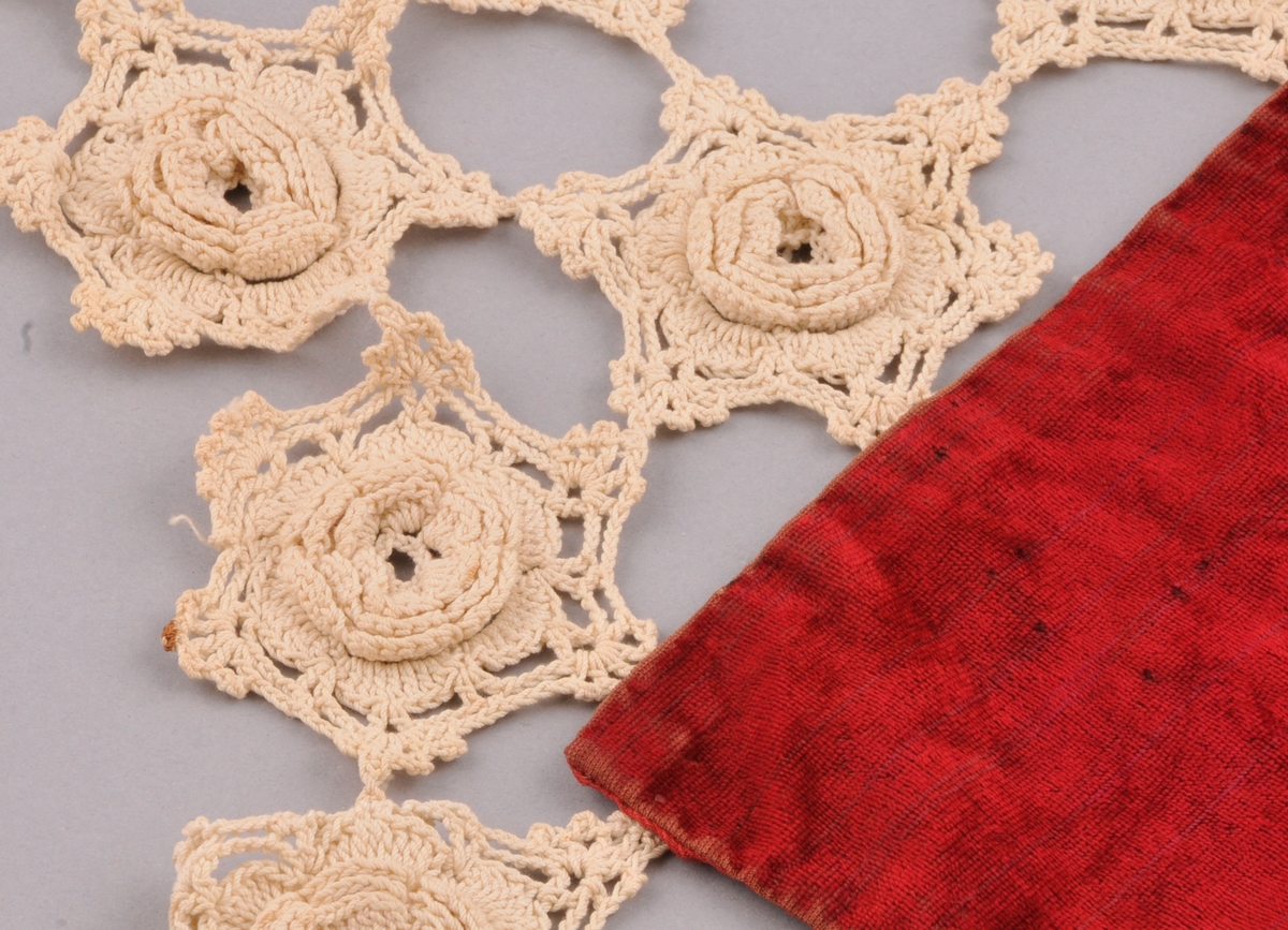 Kvadratisk tekstil sett saman av ein lapp av fløyel, med bakstykke av kypervove raudt stoff, og ei hekla blonde rundt kanten.