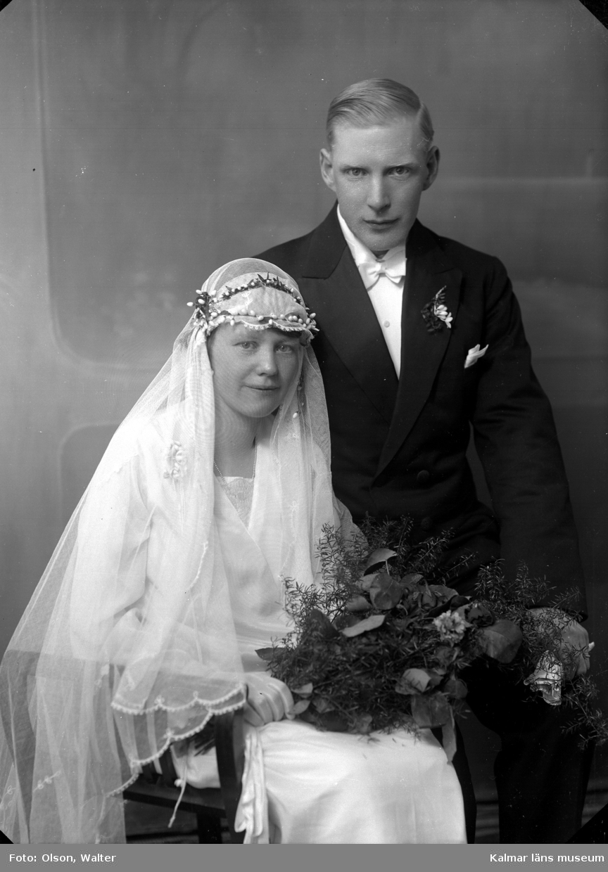 Bröllopsbild i ateljé, kvinnan har slöja och håller i en blombukett. Mannen har frack. Enligt Walter Olsons journal är bilden beställd av Henning Gustavsson ifrån Södra vägen 37 A i Kalmar.