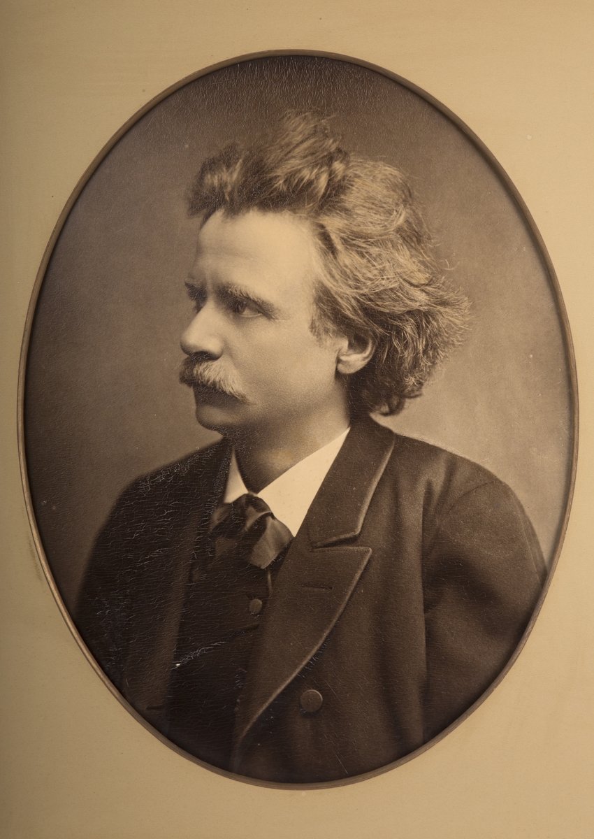 Portrettfotografi av Edvard Grieg, satt i oval passepartout som har gullkant inn mot fotografiet.