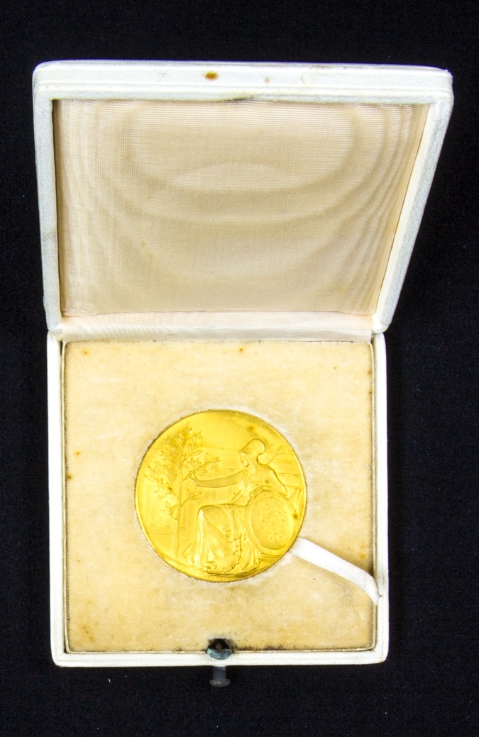 Medalj av guld med präglad text i kvadratiskt etui klätt med vitt skinn samt diplom.