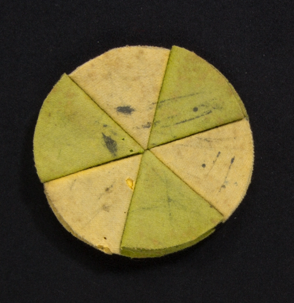 Penntorkare, rund, tillverkad av bitar av ylletyg i flera lager. Varje bit vikt i flera lager till en triangel och hopsydda med varandra. Varannan triangel grön, varannan gul och undersidan svart. Spår av användning.