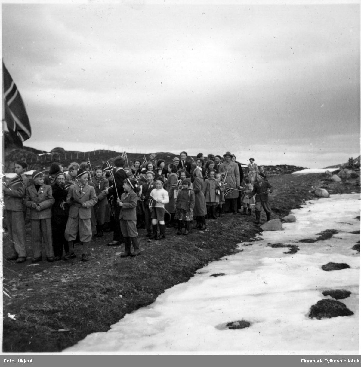 17.mai feiring i Nuvsvåg etter frigjøringen. Fotografiet tatt på slutten av 1940-tallet. Barn går i 17.mai tog med norskeflagg. Barna er kledd i skjørt, luer, bukser, gensere, jakker og sko. Man kan se enkelte menn kledd i jakker og hatter. På bakken ligger det snø.