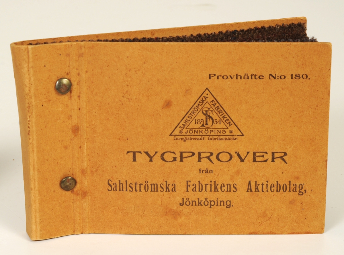 Provhäfte med tygprover från Sahlströmska Fabriken i Jönköping. Häftet innehåller 8 tygprover av mörka ylletyger. Tygproverna sitter mellan två omslag av kartong, sammanhållna av två metallklämmor.