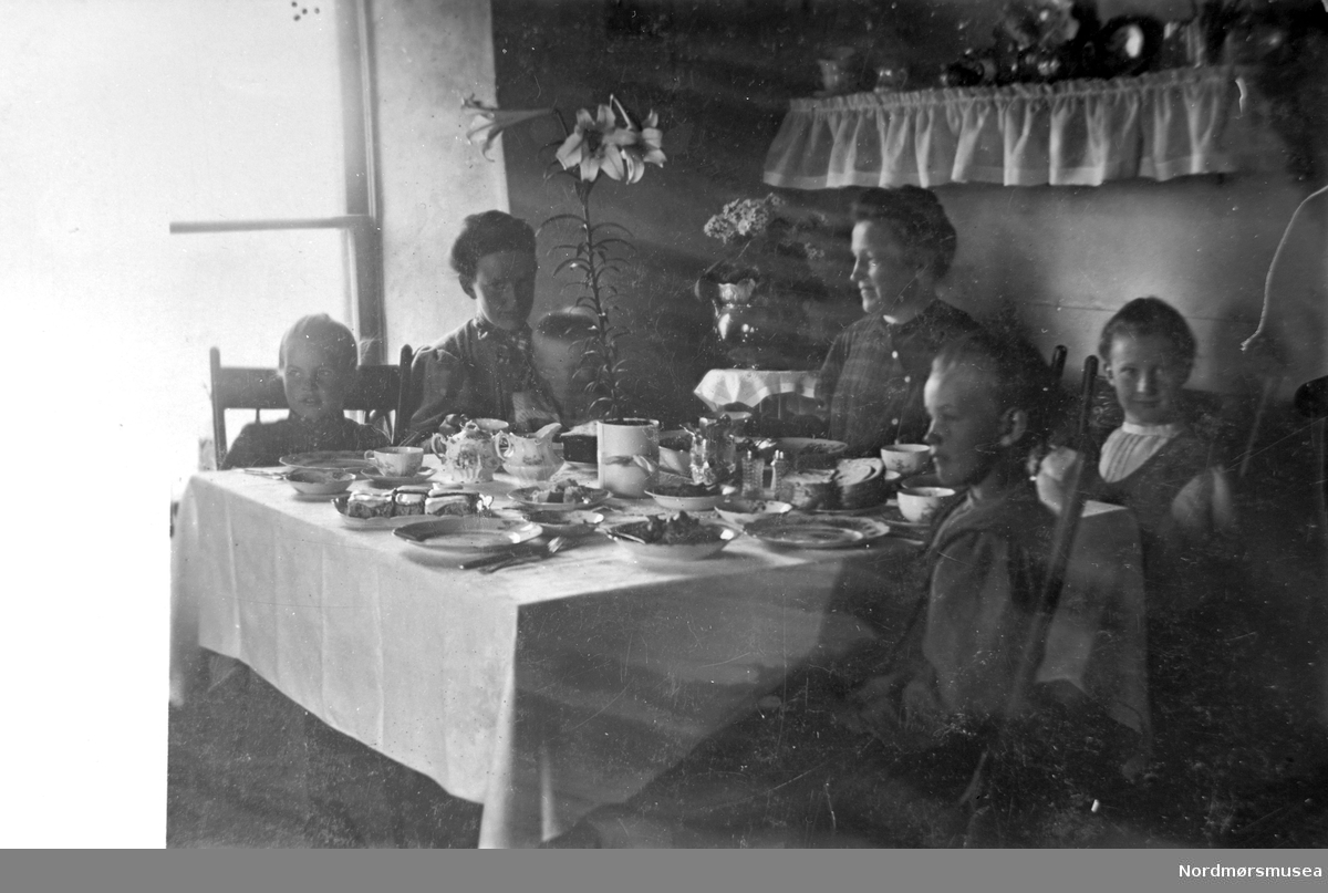 Foto av to kvinner, tre piker og en gutt sittende ved et matbord. Det er usikkert hvor og når bildet er tatt, samt hvem vi ser på bildet. Det er trolig fra Kristiansund. Fra Nordmøre Museums fotosamlinger.
