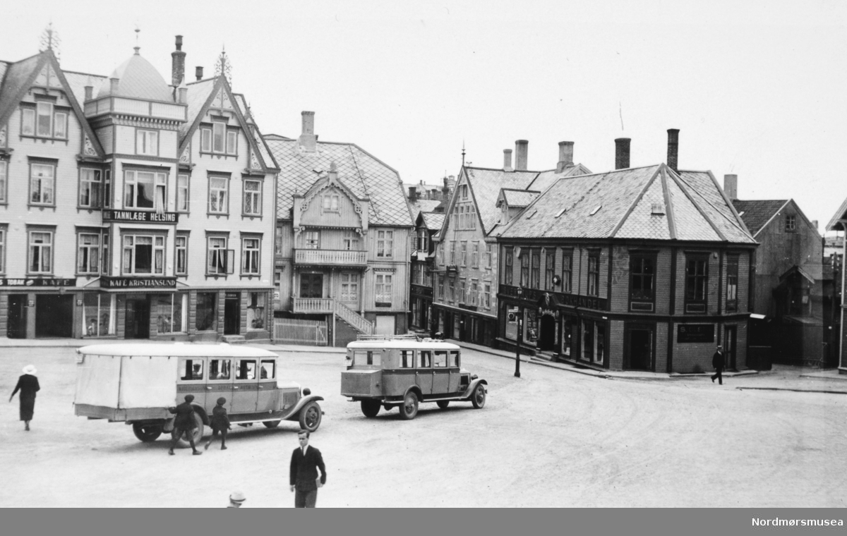 To busser står parkert på Torget på Kirkelandet i Kristiansund. Vi ser Sverdrups bokhandel til høyre i bildet. På bilen til venstre kan vi skimte Kristiansund-Oppdal Auto (KOA) på stripa under vinduene. Her er Oppdal stavet med P. Dette er nok av de tidlige rutebilene fra KOA. I 1932 åpnet ferjesambandet Kristiansund-Kvisvik. KOA og Surnadal Billag sine rutebiler kunne dermed følge ferja til Kristiansund sentrum. Det var først i 1938 at rutebilene fra Molde og Gjemnes kunne følge ferja til Kristiansund. Det var dette året ferjesambandet til Gjemnes og Torvikbukt åpnet. Bildet er derfor tidligst fra 1932. (info Sveinung Berild - 11.11.2016) Fra Nordmøre Museums fotosamling.
