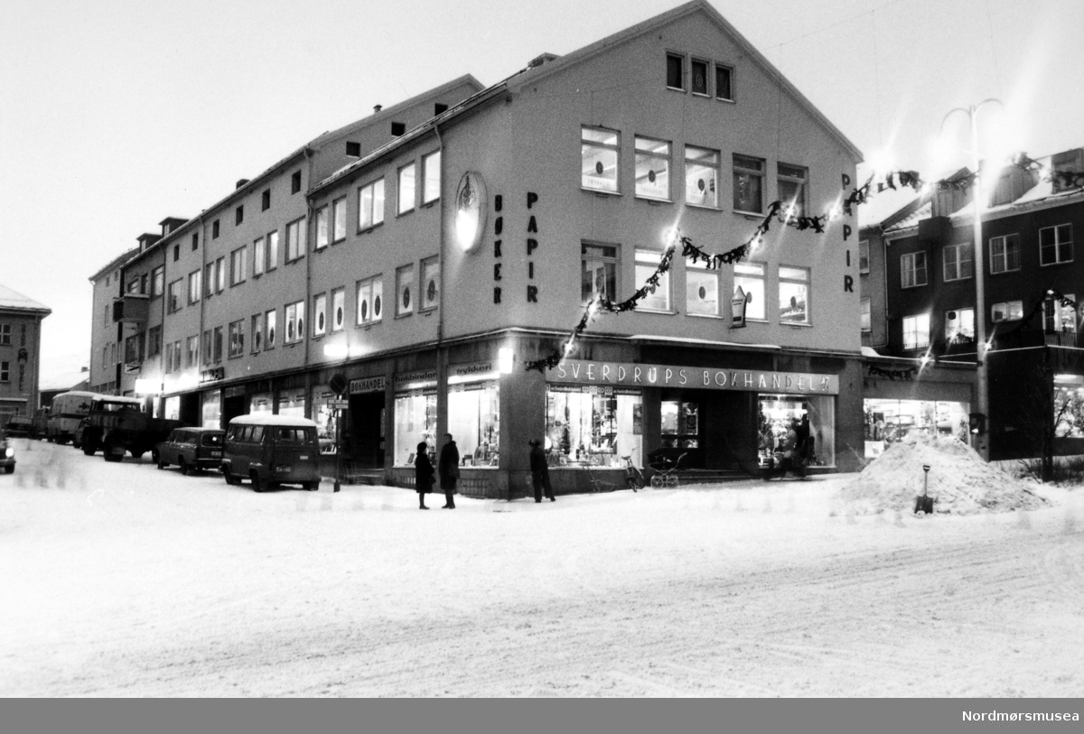 Foto fra nybygget til Sverdrups bokhandel. Det ble påbegynt rundt februar 1947, da sprengningsarbeidet var ferdig. 6. mars 1948 ble murfoten reist, mai 1948 begynte forskalingsarbeidet og 6. desember 1948 var bygget reist, og allerede høsten 1949 kunne man da starte arbeidet med å flytte inn! Firmaets samlede gulvflate var nå på hele 2550 m2 og de vanskelige arbeidsforholdene med blant annet lokaler og lager på 9 forskjellige plasser i Kristiansund var slutt. I 2012 la Ark Sverdrup ned sin drift, og nå er Den Norske Bank
(DNB)i ferd med å ta over bygget. Bildet er trolig fotografert 18. desember 1973. Fra Nordmøre Museums fotosamlinger. Kilde: Sverdrups bokhandel 100 år 1874 - 1974. Side 75-76.
