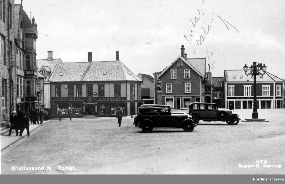 Postkort ";574"; med motiv fra torget på Kirkelandet, hvor vi ser blandt annet kommunehuset til venstre. Deretter kommer Sverdrups Bokhandel som lå i Torvgaten 9. Georg Sverdrup hadde kjøpt lokalene 1. oktober 1930, fra Fosna Mållag, som reiste bygget i 1910. Bedriften var da eid av G. Sverdrup. 28. april 1940 brant butikken ned til grunnen og den 24. september 1940 ble branntomten overført fra G. Sverdrup til Sverdrups Bokhandel A/S. Bygget i midten er Dahls bygård og inneholdte blant annet en konfeksjonsforretning, mens huset ytterst til høyre er Berseth, som inneholder blandt annet manufaktur og teppeforetning. Ellers så ser vi tre biler stående foran gatelyset på torvplassen ved kommunehuset. Utgiver av dette kortet er Georg Sverdrup og Sverdrups bokhandel, som også er fotograf. Datering er trolig omlag 1930 til 1939. Fra Nordmøre Museums fotosamlinger. Reg: EFR
