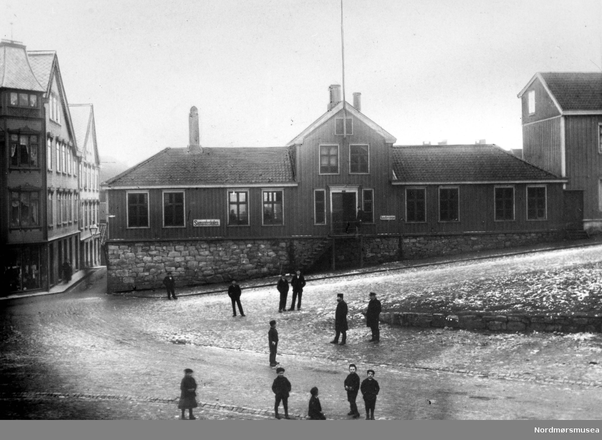 Bernstorffskolens "Sømandsskole". I 1905 ble det vedtatt å selge Bernstorffskolen med 1/2 mål grunn til Norges Bank for 15.000 kroner. Til da hadde Norges Bank holdt til i Sparebank-gården i Skolegaten, som var byens "finansgate" på den tiden. De andre bankene hadde også kontor i denne gaten. Bygningen ble tegnet av Kristen Tobiaz Rivertz fra Kristiania, som oppholdte seg da i perioden 1907 til 1909 i Kristiansund, hvor han blandt annet har tegnet "en Badeanstalt", Festiviteten, Grand Hotell og Norges banks lokaler. Norges Banks nye lokaler stod ferdig i 1909, hvor de stod frem til 28. april 1940 da banken brant ned sammen med store deler av byens bebyggelse. Da krigen var over ble det nye Norges Bank oppført i perioden i 1945-1952 og ble da tegnet av Arkitektene Eivind Moestue og Ole Lind Schistad. I dag (2007) holder Kristiansund folkebibliotek til i dette bygget. I 1915 flyttet Tidens Krav inn her i samme lokaler med sitt trykkeri. Her holdt de til inntil økonomien ble for trang, og i 1924 flyttet de ut av lokalene og inn i nabobygget - Arbeiderforeningen. 
Fra Nordmøre Museums fotosamlinger.
Kilde (Bl.a): Eivind Aass: Kristiansunds Historie VI, side 43
Kilde: Petter E. Innvik: Et fyrtårn i havgapet. Tidens Krav gjennom 100 år. 1906 - 2006, side 41 og 47. EFR2015