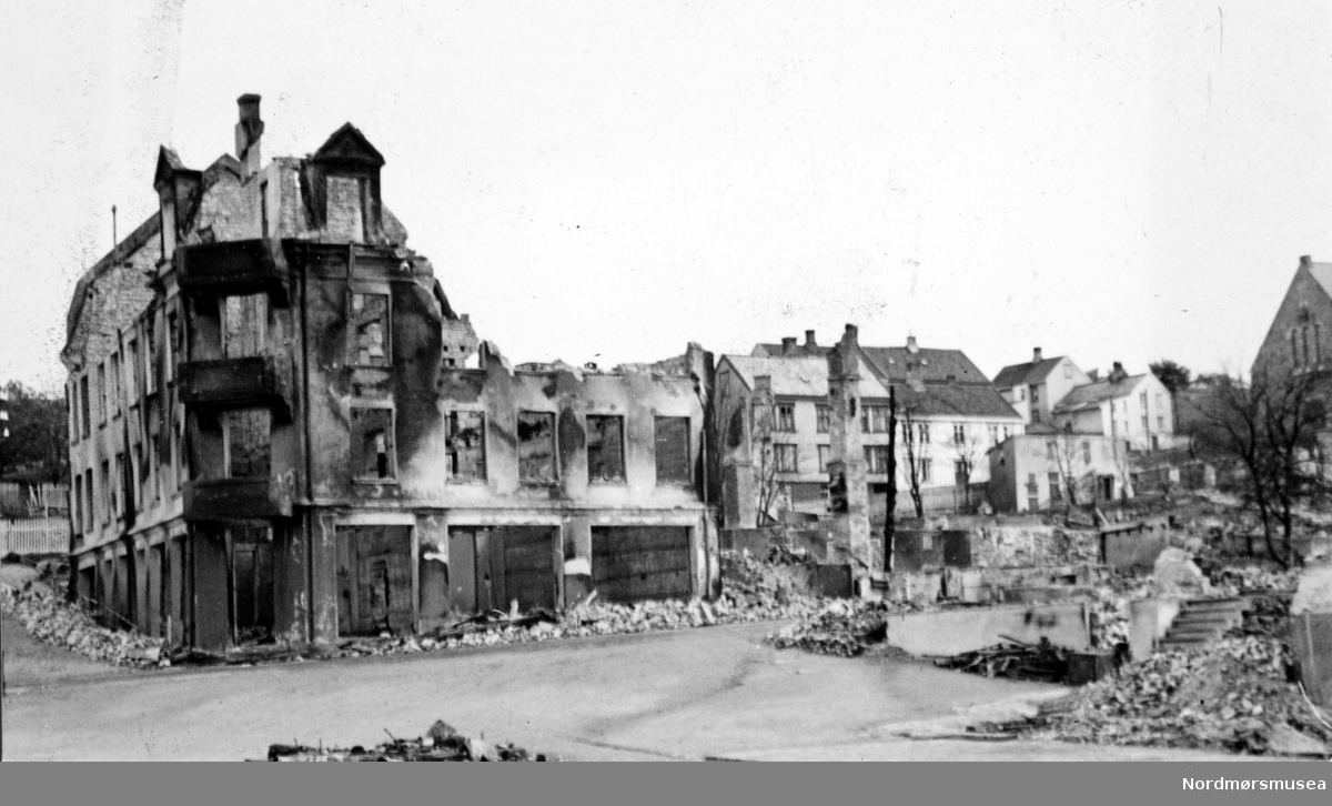 Krigen har kommet til Kristiansund. Etter nazistenes herjinger i perioden 28. april til 1. mai 1940 ligger byen i ruiner. Det totale antall brente bygg var 767, hvor 605 av disse var på Kirkelandet og 162 på andre "land" i Kristiansund. Her fantes det til blant annet til sammen 3906 boliger ifølge boligtellingen av 1938, og av disse brant 2162 boliger ned og 7099 mennesker ble husløse. I tillegg til de 767 brente byggene brant 36 fiskepakkehus ned til grunnen, hvorav 34 lå på andre "land". Verdien av de brente byggene beløp seg til kroner 26,9 millioner for Kirkelandet (kroneverdi per 1940) eller tilsammen 30,6 millioner kroner (kroneverdi per 1940) for hele byen. Fra Nordmøre Museums fotosamlinger. Kilde: Gjenreisningsproblemer i Kristiansund. Fremlagt ved gjenreisningsinstituttet i Kristiansund. Juli 1945. Side 16 - 20. EFR2015