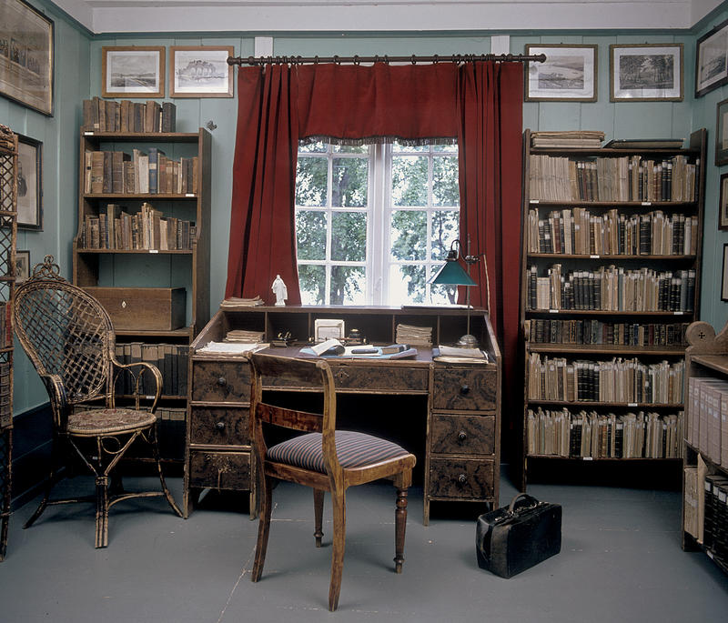 Interiør; bibliotek med bokhyller på begge sider av et vindu med røde gardiner, skrivebord og stol foran vinduet. (Foto/Photo)