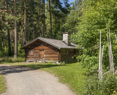 Brunt tømmerhus med flistak og høy pipe ligger idyllisk langs en grusveg med en skigard til høyre og høyre furuer bak.