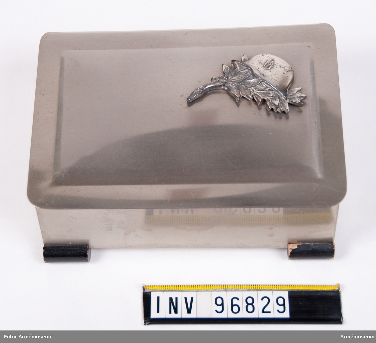 Skrin i metall med träinredning som möjligen har använts för att förvara cigarrer. Ovanpå locket en utsmyckning i form av en blomstergirlang med en tysk hjälm.