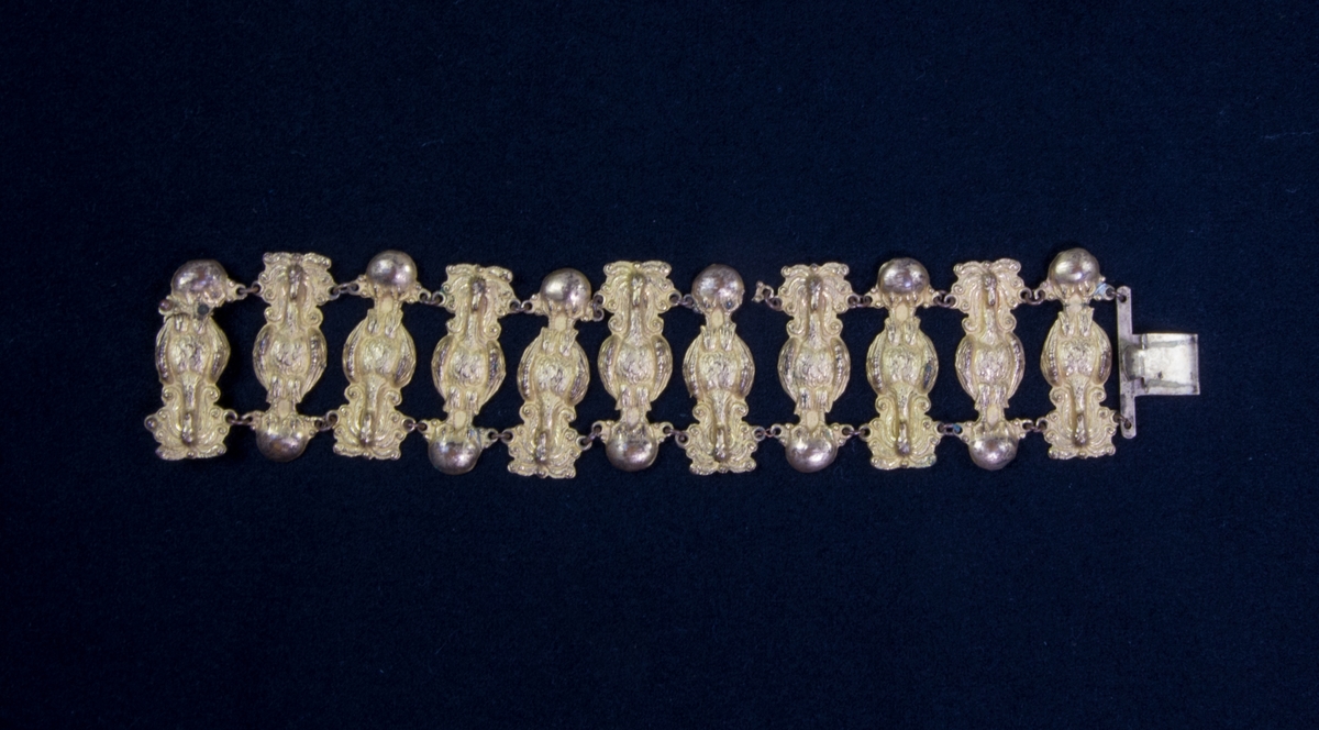 Armband av pressad mässingsplåt. Består av 11 stavliknande ornament sammansatt med kedjor. Lås.