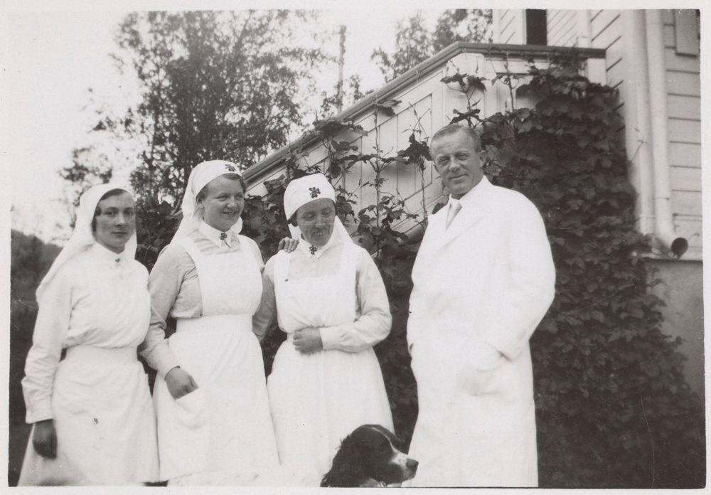 Tre sykesøstre og dr. Brochmann utenfor Gullhaugen. Hodet på en hund stikker opp i forgrunnen.