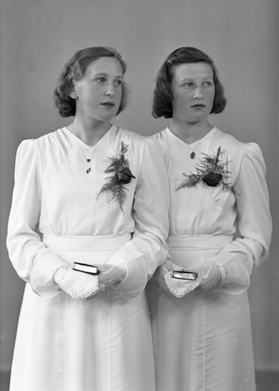 Foto av två systrar i vita konfirmationsklänningar. 
I händerna håller de små psalmböcker.
Knäbild. Ateljéfoto.
