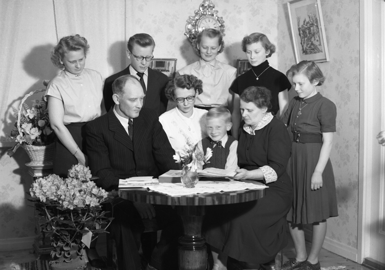 Foto av en man i kostym som sitter i ett vardagsrum. omgiven av sin familj. Man studerar en bok ("Sveriges Bebyggelse" ?) på ett bord.
