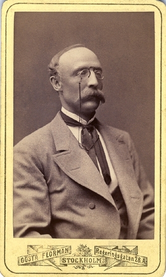 Porträtt (bröstbild, halvprofil) av en okänd man med mustasch och pincené, klädd i ljus kavaj med väst 
och vit skjorta med stärkkrage och mörk slips/spännhalsduk.