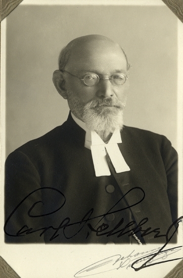 Porträtt (bröstbild, halvprofil) av en äldre präst i prästdräkt, skägg och glasögon.
Snett över fotografiet finns personens autograf: "Carl Hellberg."