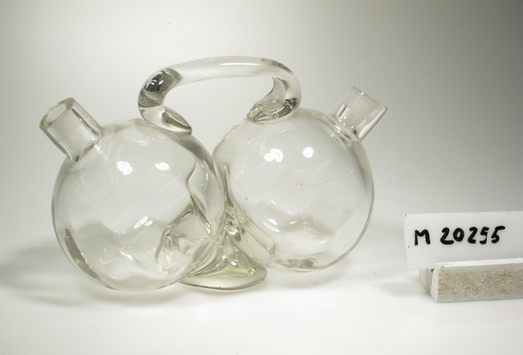 Dubbelflaska med grep.
Tillverkad på Sandvik-Orrefors glasbruk 1920.
Två små flaskor som är ihopsatta med en gemensam grep.
Optik.
Klarglas.

Gåva av Orrefors glasbruk år 1968.
Funktion: Dubbelflaska (för olja/vinäger ?)