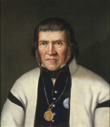 Portrett av Tallev O. Huvestad. Hvit trøye (jakke) og mørk vest kantet med rødt. Medalje hengende i kjede rundt halsen.