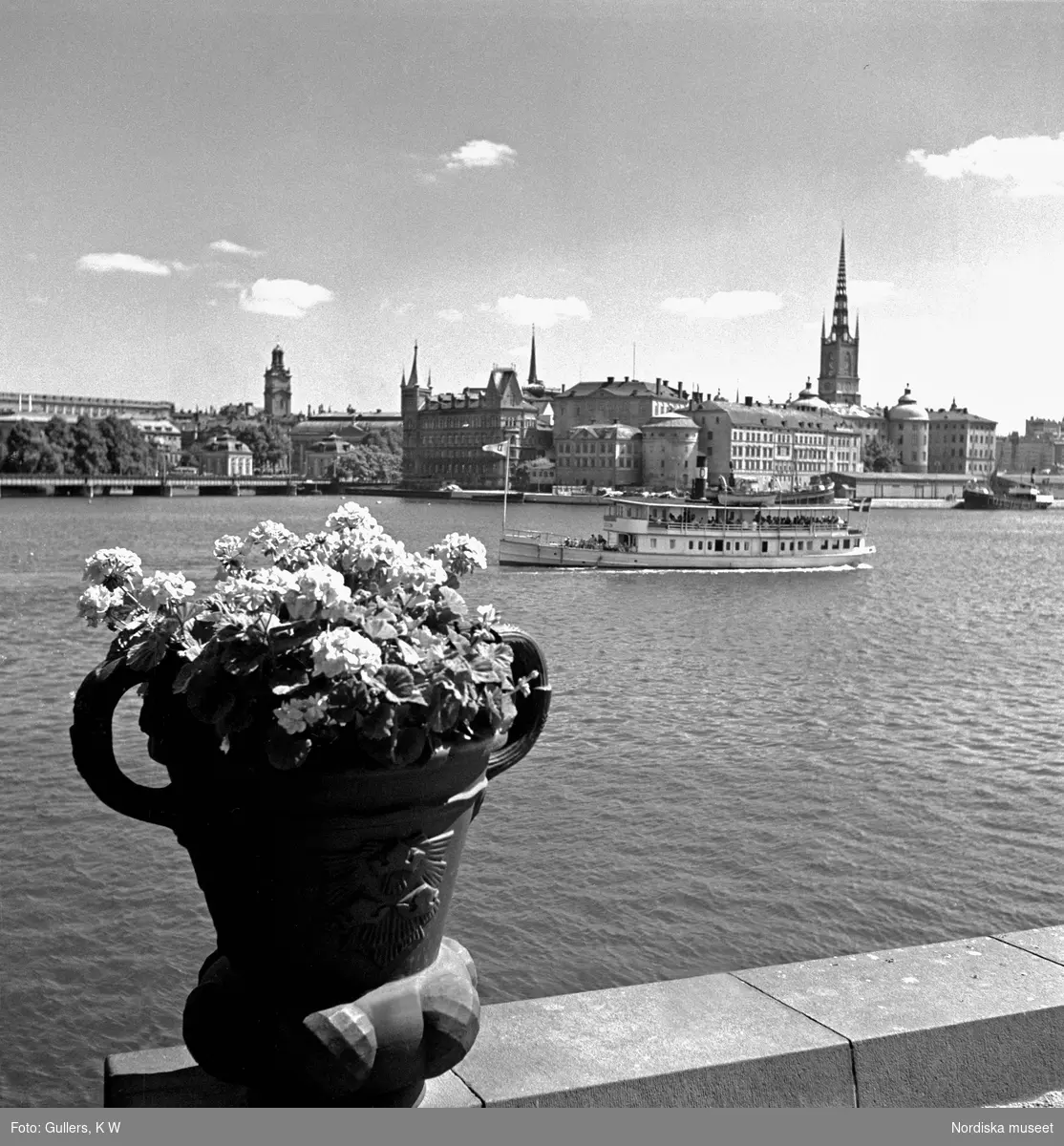 Vy från Stockholms stadshus. Skärgårdsbåt på Riddarfjärden, en blomsterurna i förgrunden, Riddarholmen och Gamla stan i bakgrunden