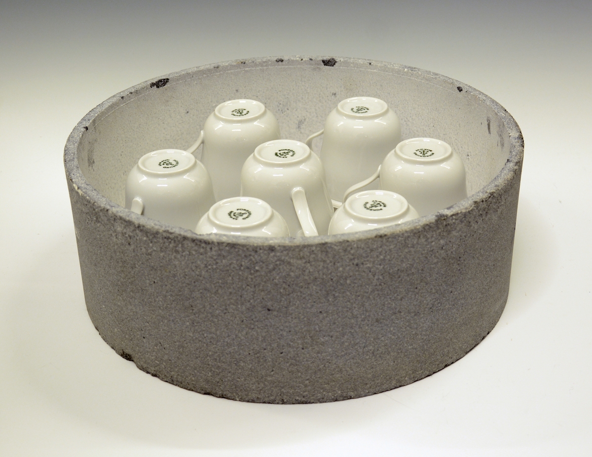 Rund kapsel i ildfast stein med syv porselenskopper på bumser/styringer (Merket A-G).