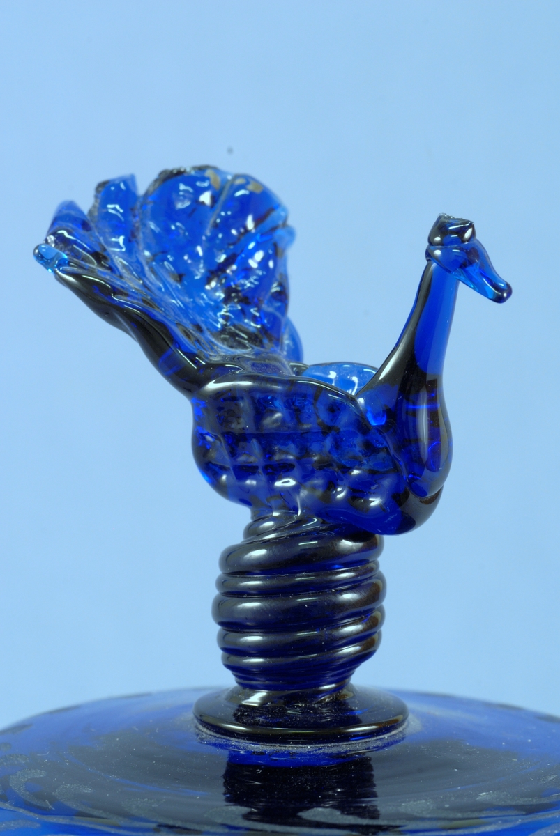 Lockskål med två hankar av blått glas. Lockknopp av fritt modellerad fågel. Graverad dekor med initialerna NAS och MLJD samt en blomma på skålen. Bård av ax och kvistar med mittblomma. På locket en enkel bladbård.