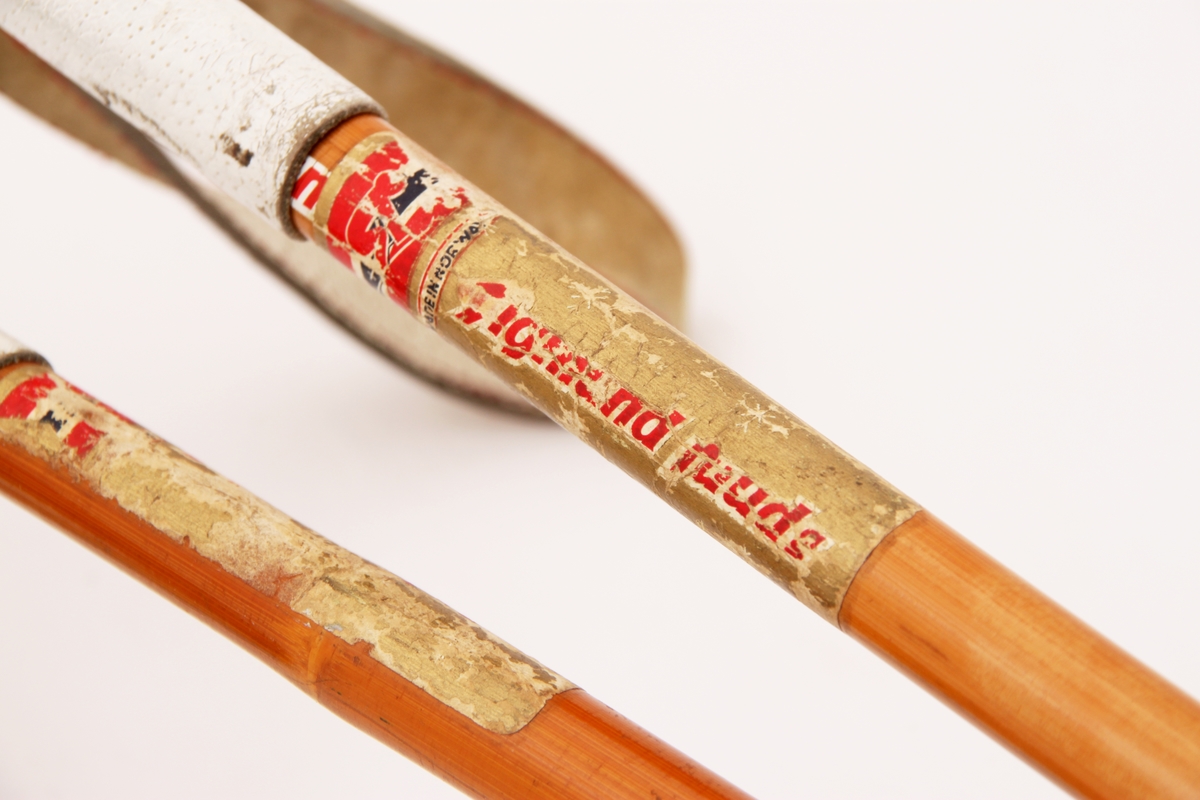 Et par bambusstaver med spanskrør/rotting-trinse med røde lærkryss. Stavene har påsydde lærhåndtak og sydde håndstropper i hvit farge.