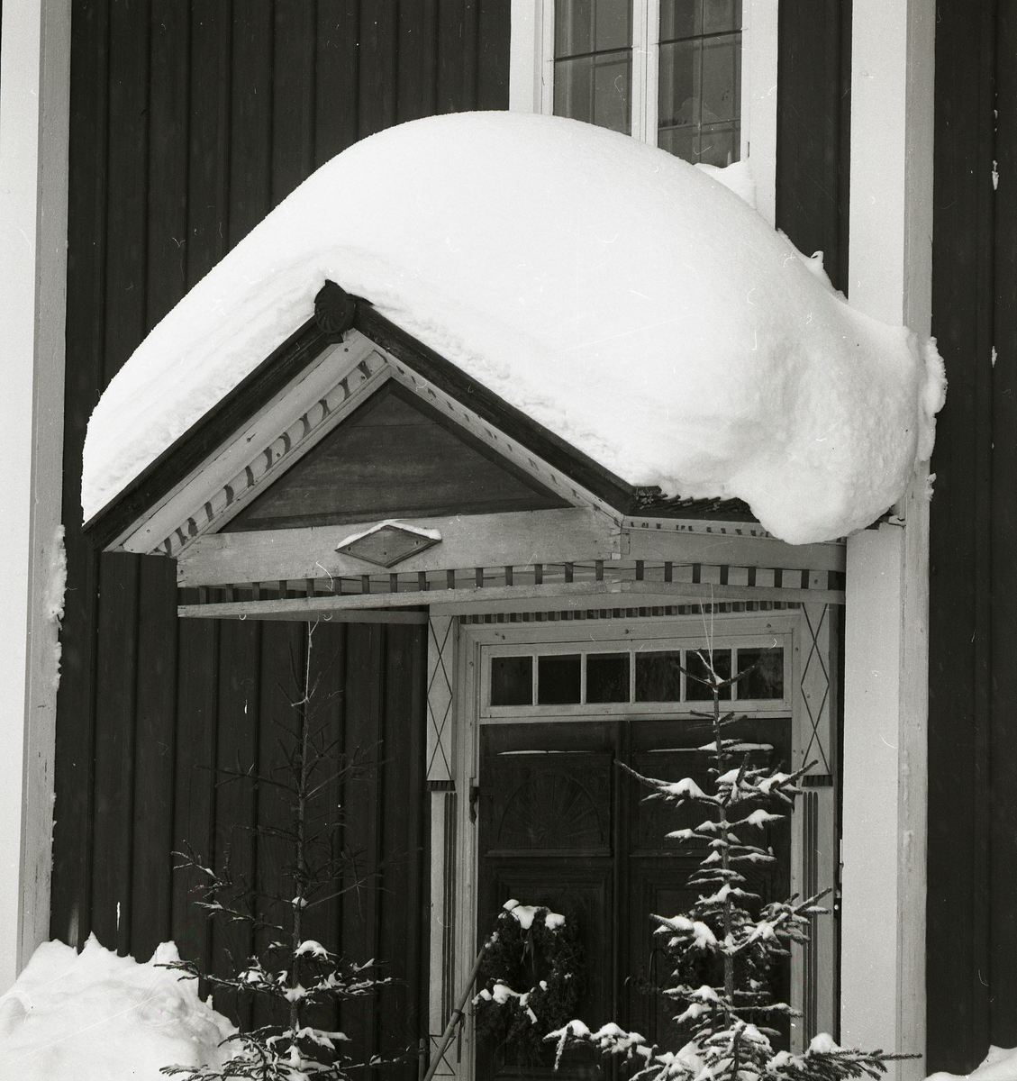 Tung ligger snön på farstukvistens tak och når ändå upp till fönstrets undre kant. På pardörrarna under farstukvisten hänger grankransar och berättar att julen är i antågande. 
Två granar ramar in dörren på sidorna och överljusfönstret markerar dörrens avgränsning upptill.