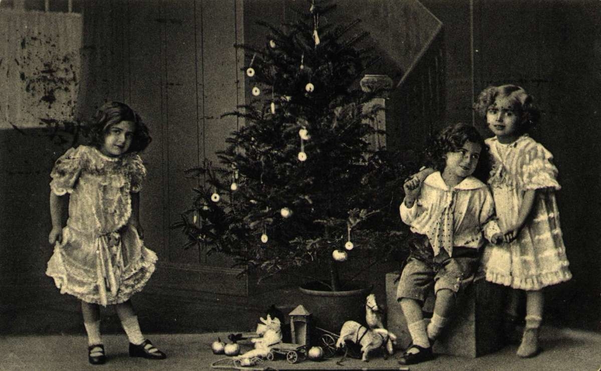Julekort. Jule- og nyttårshilsen. Sort/hvitt fotografi. Festkledde barn står ved juletreet. Gaver ligger under treet. Stemplet 23.12.1911.