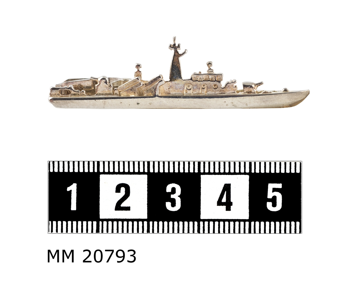 Silveraktig metall som föreställer en kustkorvett, Göteborgsklass, sedd från styrbordssidan. På baksidan av båten finns en tandad klämma. Under klämman är det två fördjupningar där det står "JOE" och "925".