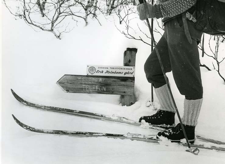 En skidåkare med ryggsäck har stannat vid en skylt där snön är så djup att den nästan döljer texten, 4 april 1966. Där står Erik Holmboms gård, Svenska turistföreningen.