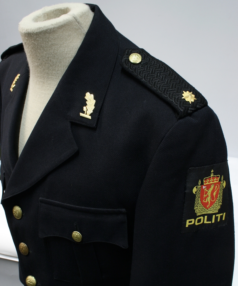 Enkeltkneppet jakke med fire riksløveknapper, fire ytterlommer, ekeblad av metall på kragespeil, skulderklaffer med en stjerne, og politimerke på ermet.