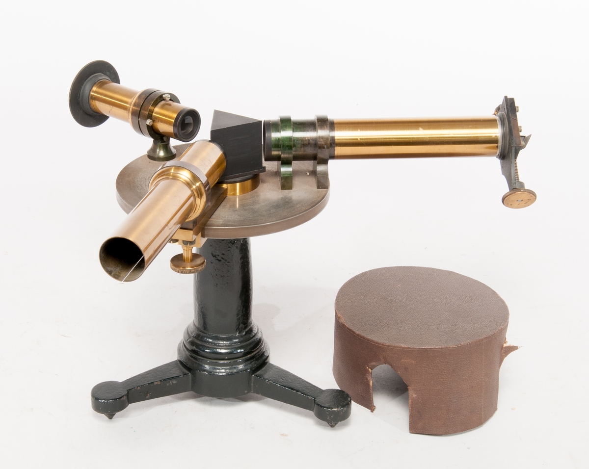 Spektroskop på fot av gjutjärn, med prisma och tuber av mässing. Skyddshuv av papp, med notering på insidan i blyerts: John Hertzberg.