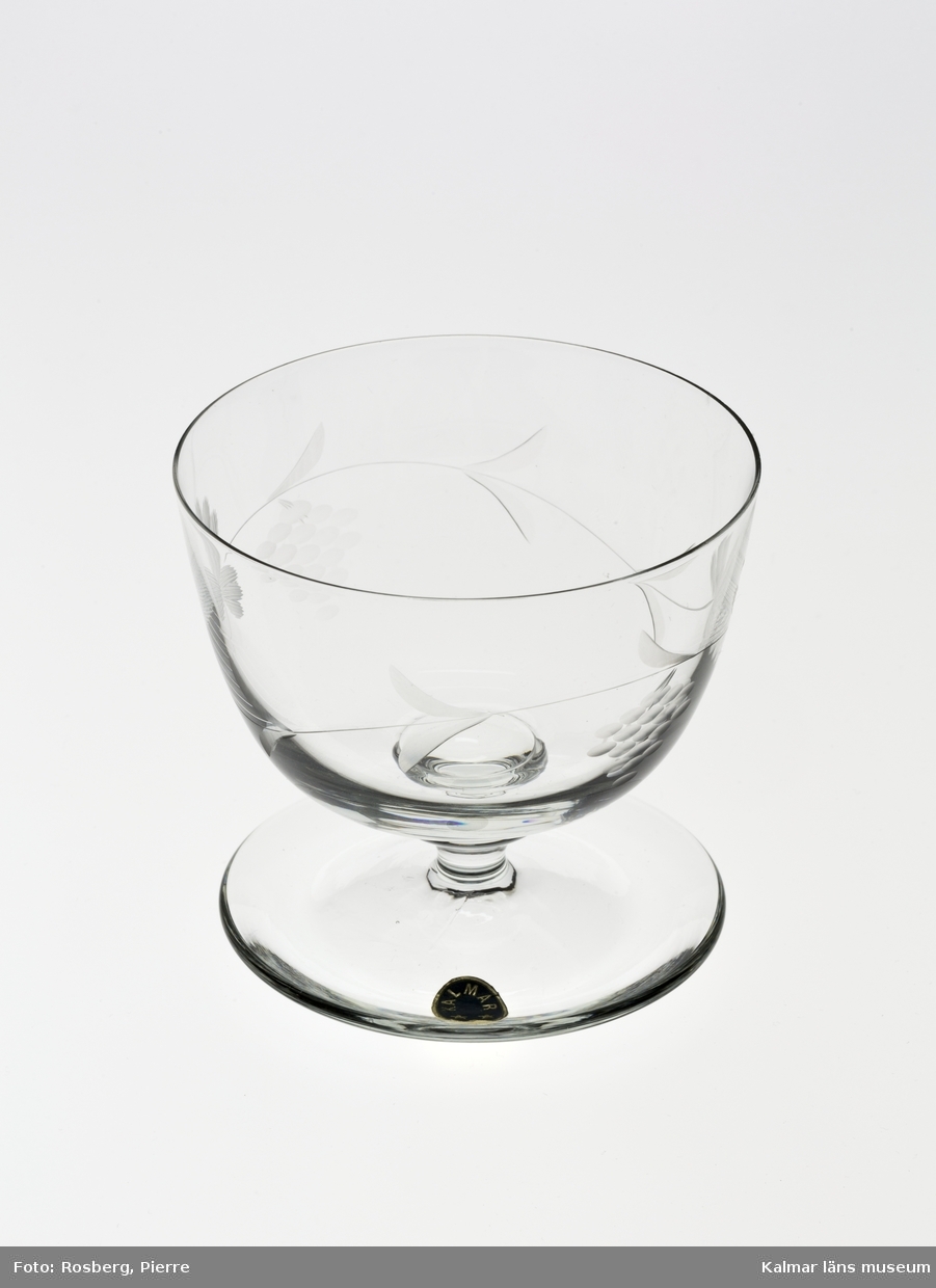 KLM 45817:1-2 Glas, dricksglas på fot. Med slipad dekor, i form av blad och druvklasar. Försedd med etikett, oval, blå botten text i silver: Kalmar och två stjärnor.
