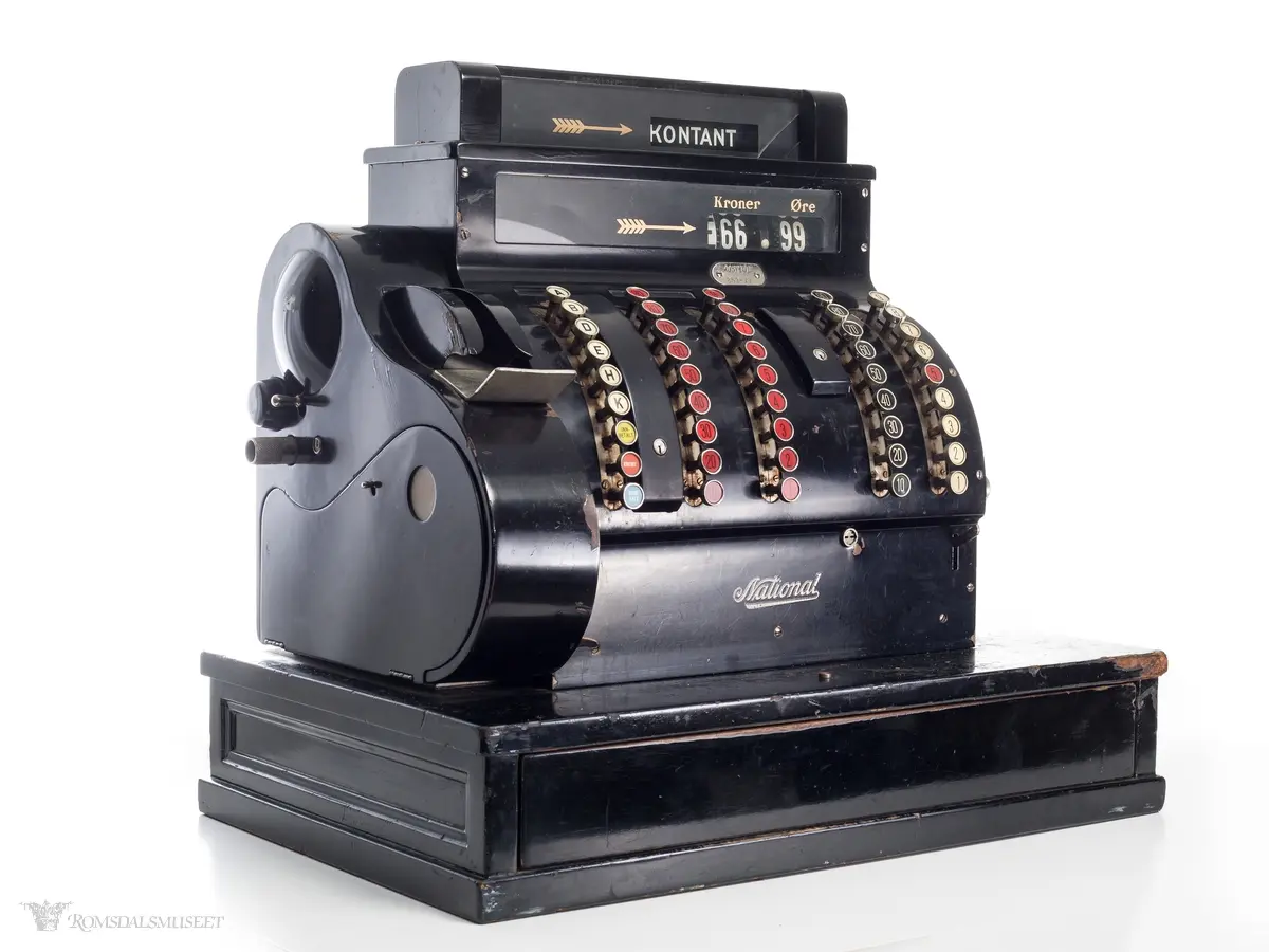 Sortfarget kassaapparat av merke National. Et kontantfelt øverst på apparatet med et felt under for tresifret kronebeløp og tosifret øre. Tre mindre felter over taster merket "KKK", "Kroner" og "Øre".

Fem rader med knapper i front. 
Raden lengst til venstre har bokstaver A-K, samt knapper for "innbetaling", "kreditt" og "utbetaling"
To tasterader på venste side med tall fra 1-9 og 10-90, og tilsvarende på høyre side.
Boks for utskrift av kvittering på vesntre side(?)

Sveiv og nøkler i lås på høyre side og av og på skru på venste side.