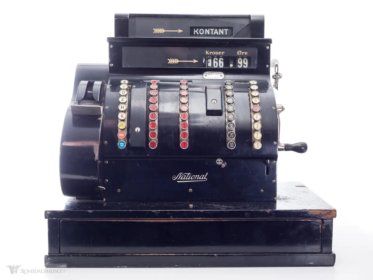 Sortfarget kassaapparat av merke National. Et kontantfelt øverst på apparatet med et felt under for tresifret kronebeløp og tosifret øre. Tre mindre felter over taster merket "KKK", "Kroner" og "Øre".

Fem rader med knapper i front. 
Raden lengst til venstre har bokstaver A-K, samt knapper for "innbetaling", "kreditt" og "utbetaling"
To tasterader på venste side med tall fra 1-9 og 10-90, og tilsvarende på høyre side.
Boks for utskrift av kvittering på vesntre side(?)

Sveiv og nøkler i lås på høyre side og av og på skru på venste side.