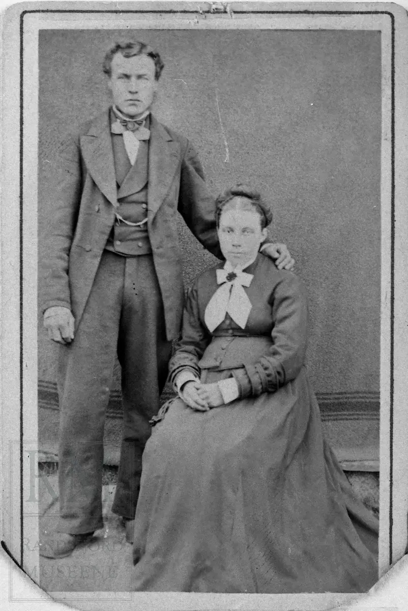 En mann stående ved siden av en sittende kvinne, han har armen på hennes skulder.