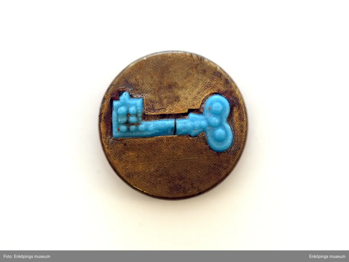 Rund knapp av järn klädd med mässingbleck. Infälld i mässingblecket en 1,8cm lång blå nyckel i ett obetsämbart material. Enligt katalogen är knappen funnen på Munksundet och är troligen från 1700- talet.