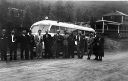 Menn og kvinner foran en buss. Skog og bygninger i bakgrunne