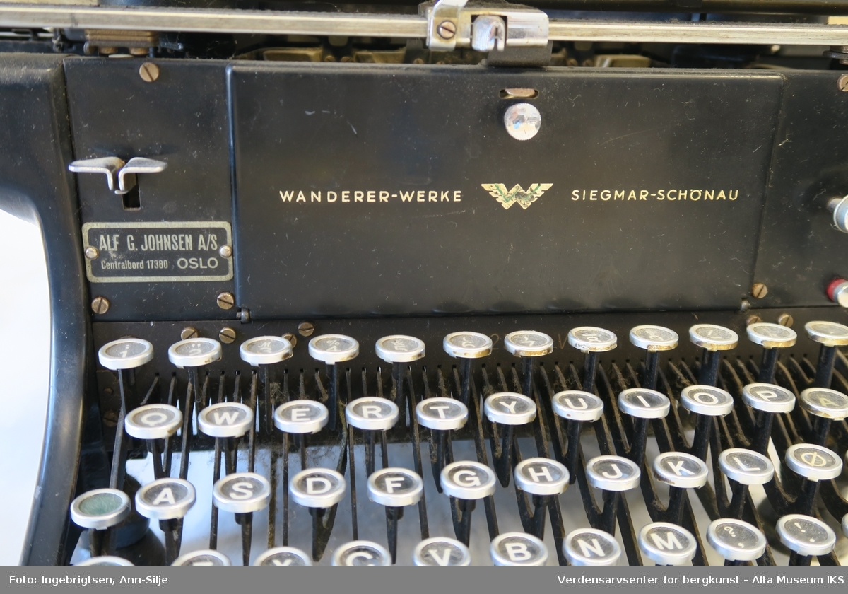 Rektangulær svartlakkert mekanisk skrivemaskin på fire bein med en bred skrivevalse som går langt utover maskinens grunnform. Maskinen har et overtrekk i tekstil.